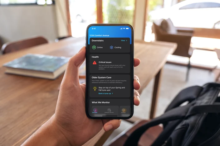 A person holds a smartphone displaying a home monitoring app. The screen shows options like online status, cooling, health alerts, system care, and monitoring features. The background includes a table, a book, and a window with a view outside.