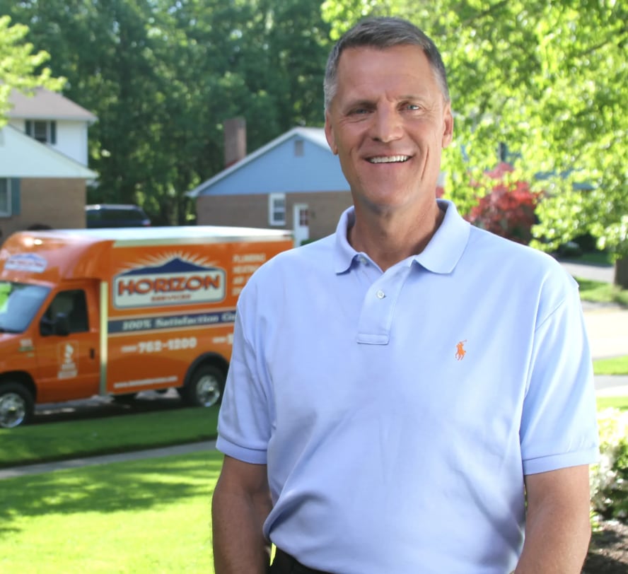 A man in a light blue polo shirt smiles while standing outdoors. In the background, there is an orange van with the Horizon logo parked on a residential street surrounded by trees and houses.