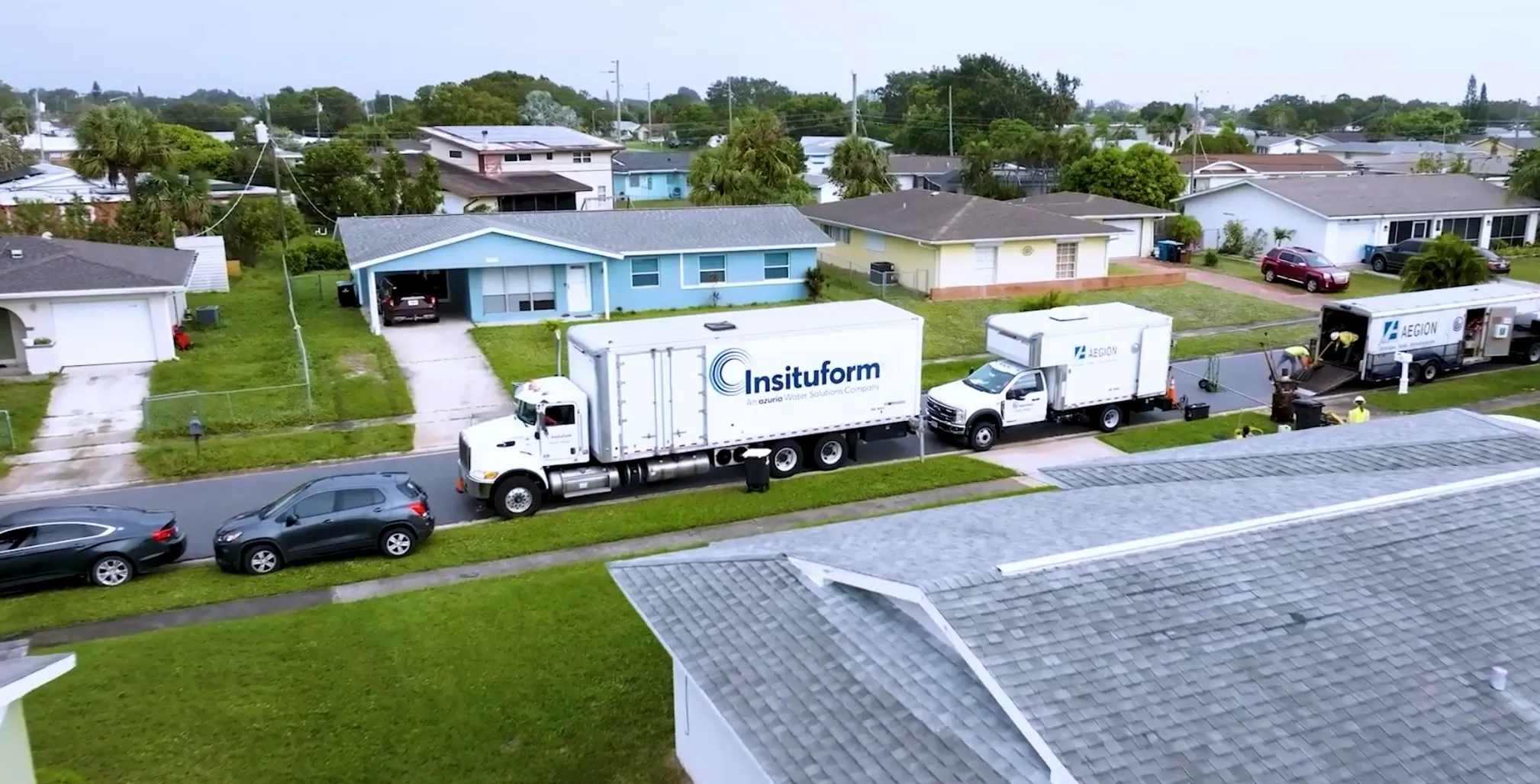 Aerial view of a residential street featuring two large white trucks with the Insituform logo parked on the road. The street is lined with single-story houses and green lawns. A gray car is parked in front of one of the trucks.