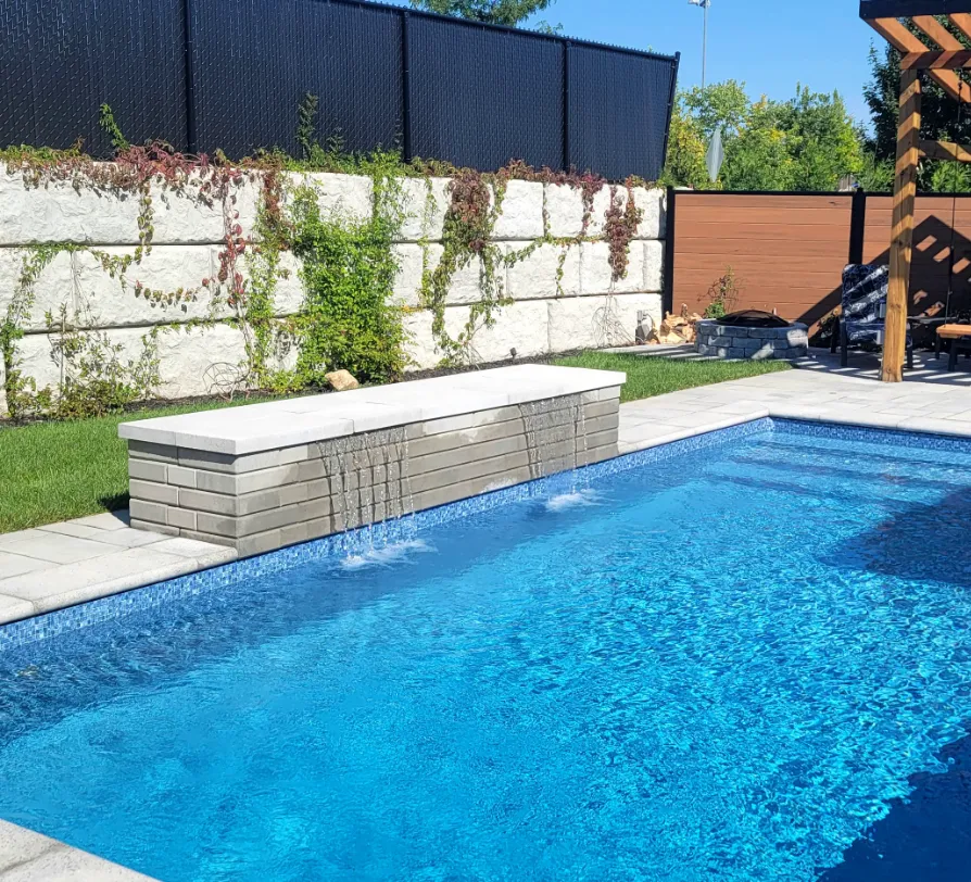 A clear blue rectangular pool with small fountains along a stone wall, surrounded by a landscaped area with grass and climbing plants. A wooden pergola is partially visible on the right side.