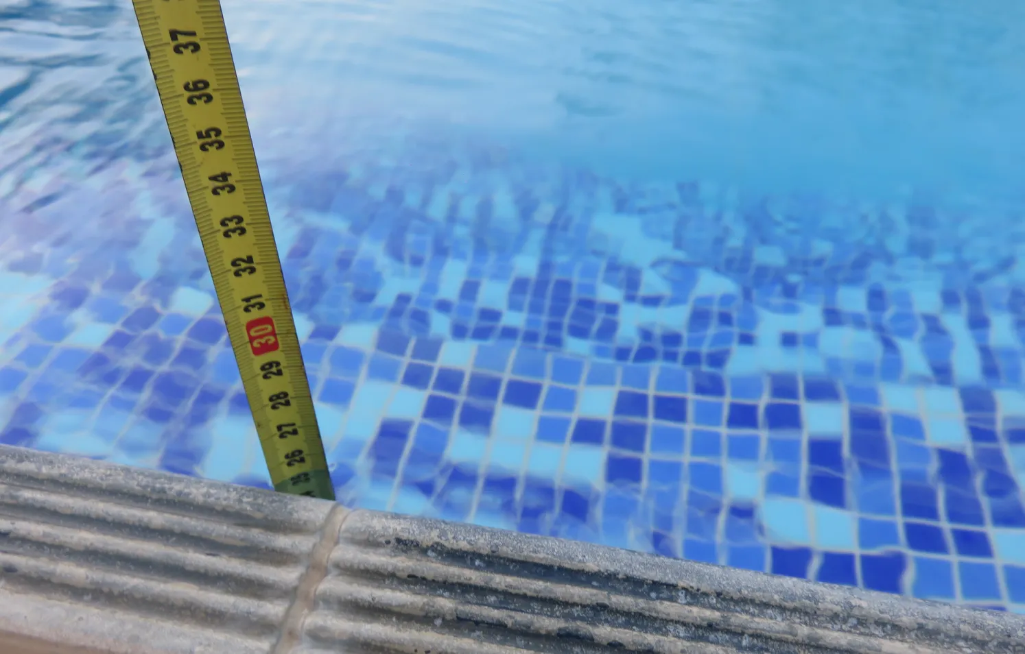 A tape measure stands vertically in a swimming pool, indicating a depth of about 36 inches. The pool has blue and white mosaic tiles under clear water. The edge features textured stonework.