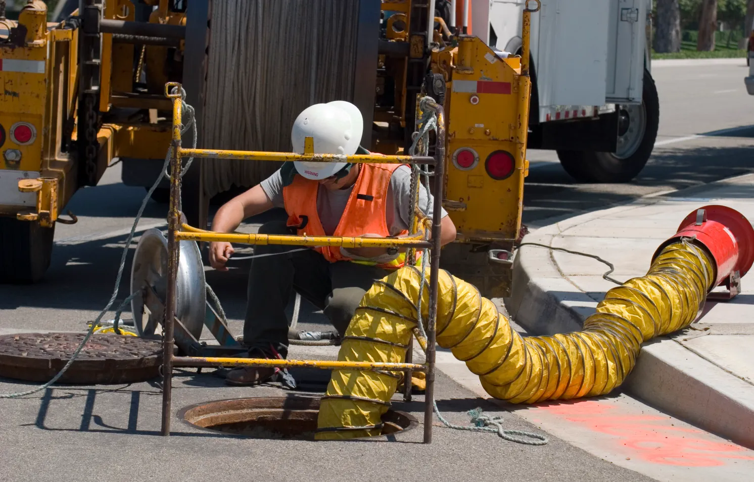 Worker wearing a white helmet and orange vest entering a manhole next to a large vehicle. A yellow flexible duct is attached, running into the manhole, with equipment set up around the opening. The scene takes place on a street with marked pavement.