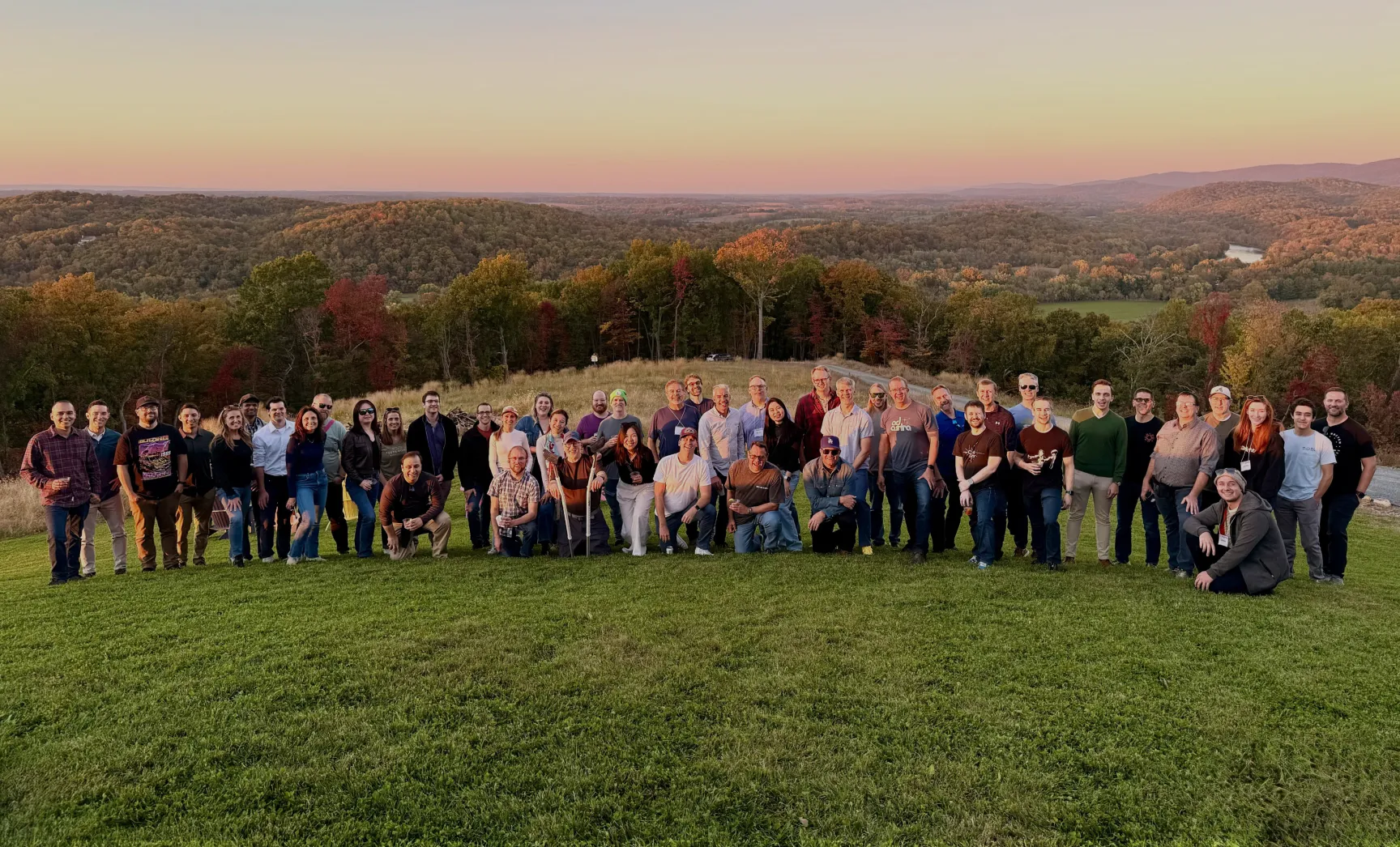 A large group of people poses for a photo on a grassy hilltop at sunset. The background features a panoramic view of distant forests and rolling hills under a colorful sky. The group is casually dressed and appears to be enjoying the scenery.