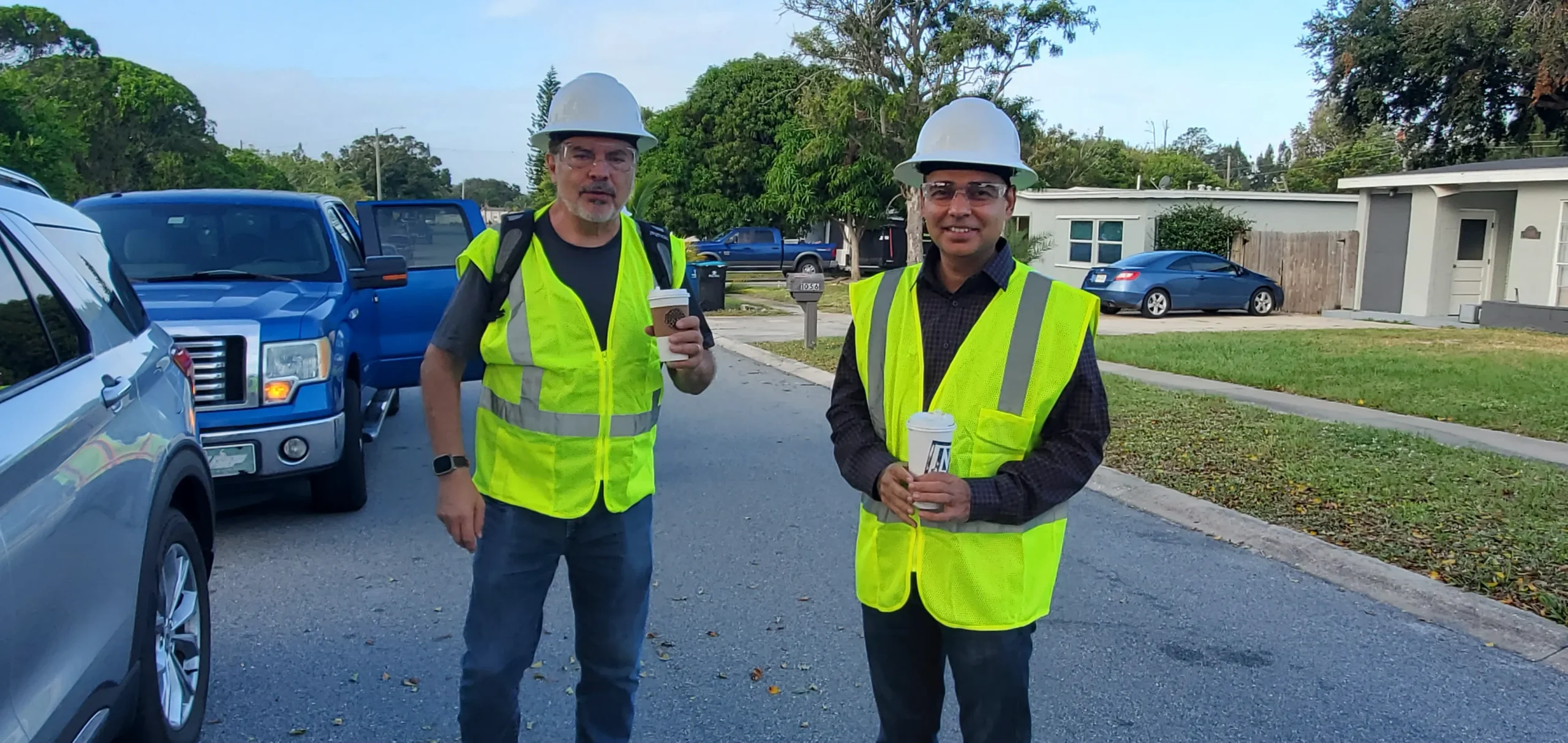 Two men in yellow safety vests and white hard hats stand on a residential street. They are holding drinks and standing near cars and houses, with trees and a blue sky in the background.