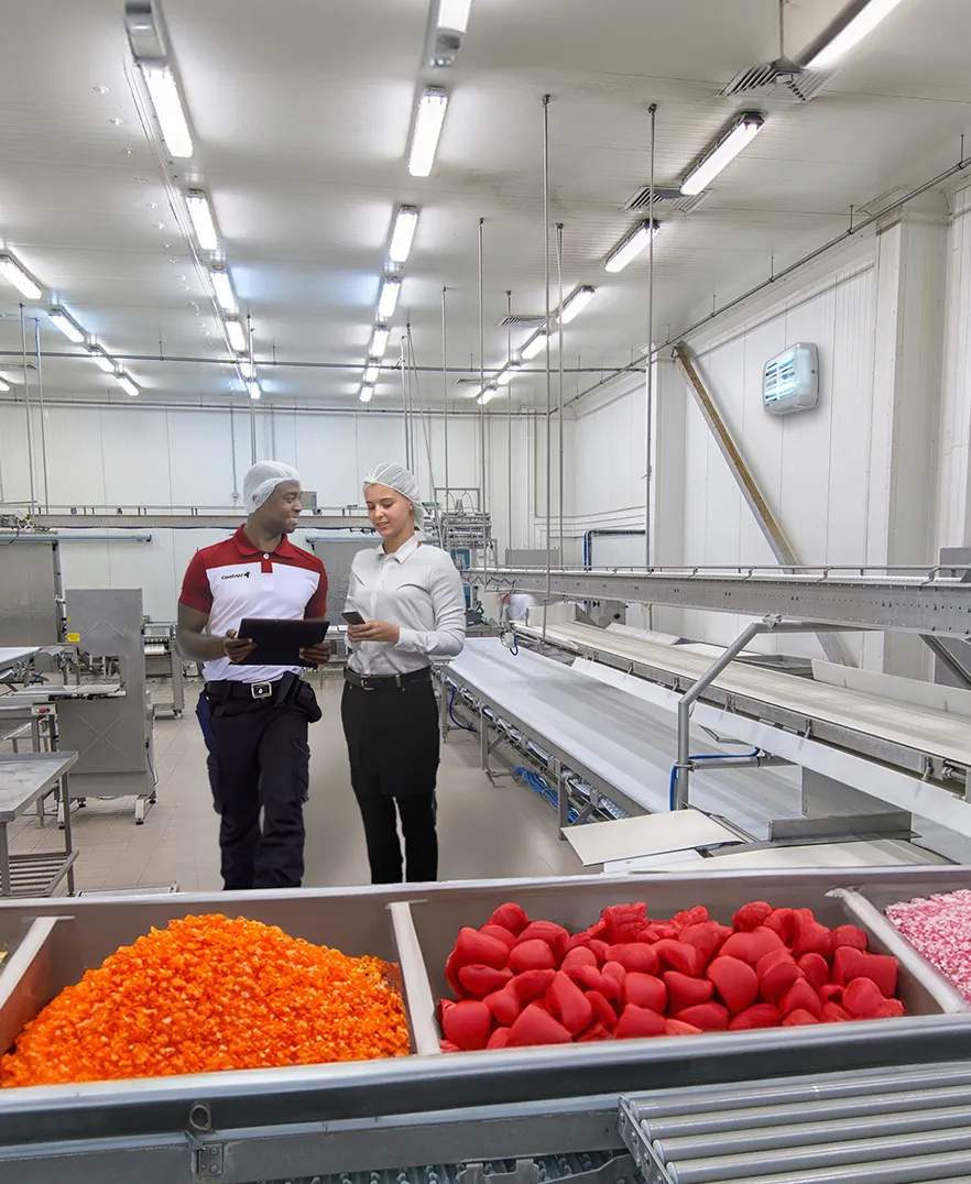 Two people in hairnets and uniforms walk through a factory with stainless steel equipment and conveyor belts. They hold clipboards and discuss in a bright room with colorful candy in bins in the foreground.